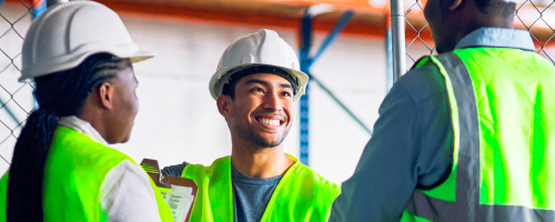 Trois ouvriers portant des gilets de sécurité et des casques de chantier sourient dans un entrepôt